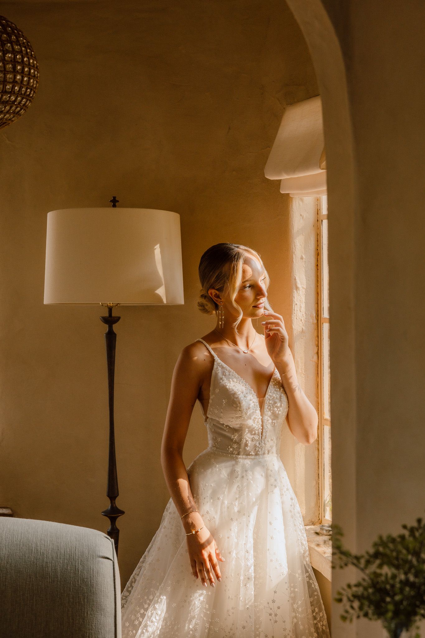 A bride in a white lace wedding dress stands in soft window light creating a romantic atmospheric moment.