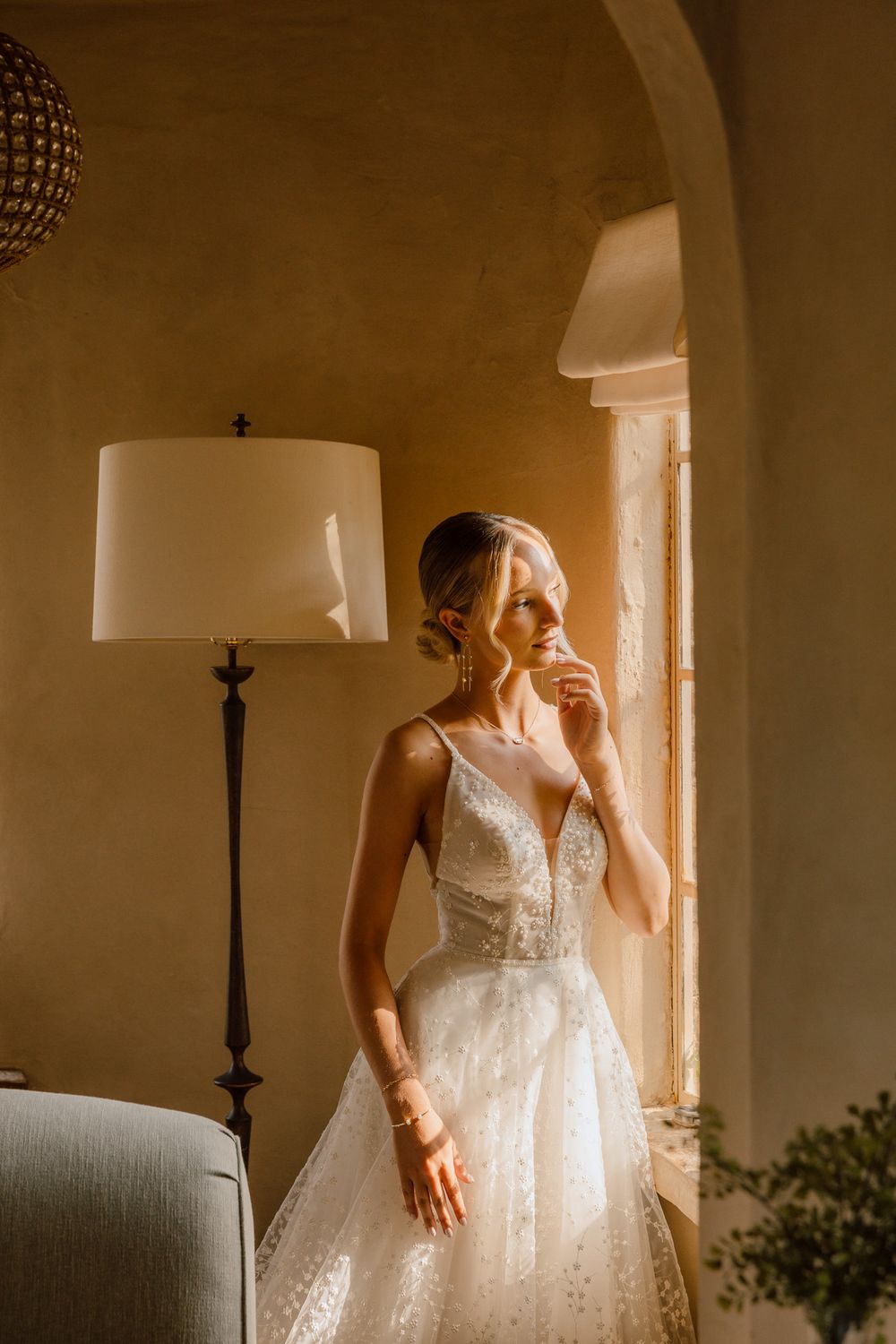 A bride in a white lace wedding dress stands in soft window light creating a romantic atmospheric moment.