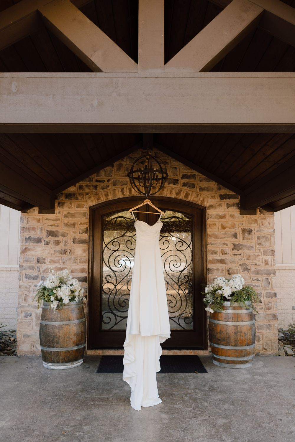 A white wedding dress hangs elegantly between rustic wooden barrels and ornate iron gates in a stone-walled venue.