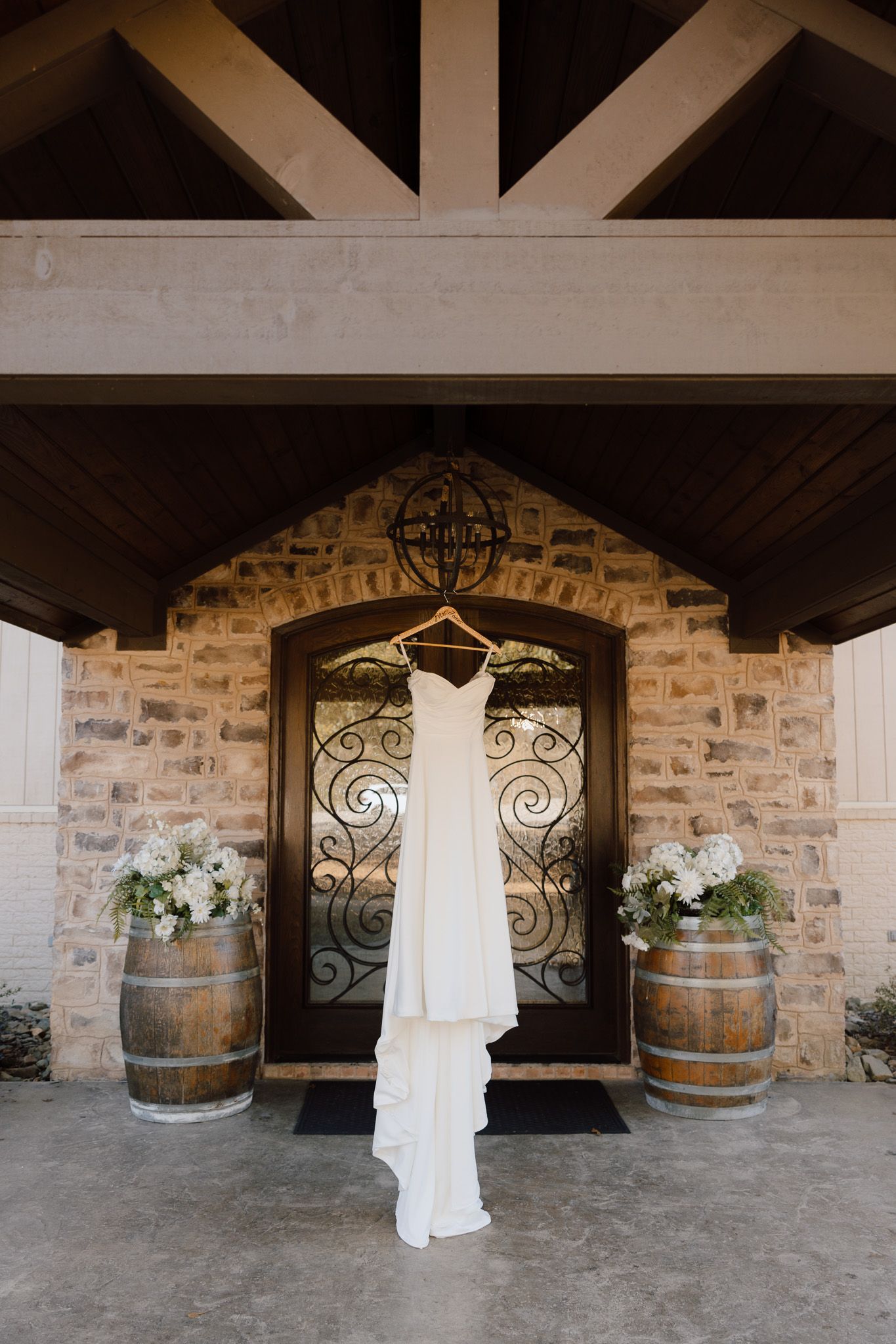 A white wedding dress hangs elegantly between rustic wooden barrels and ornate iron gates in a stone-walled venue.