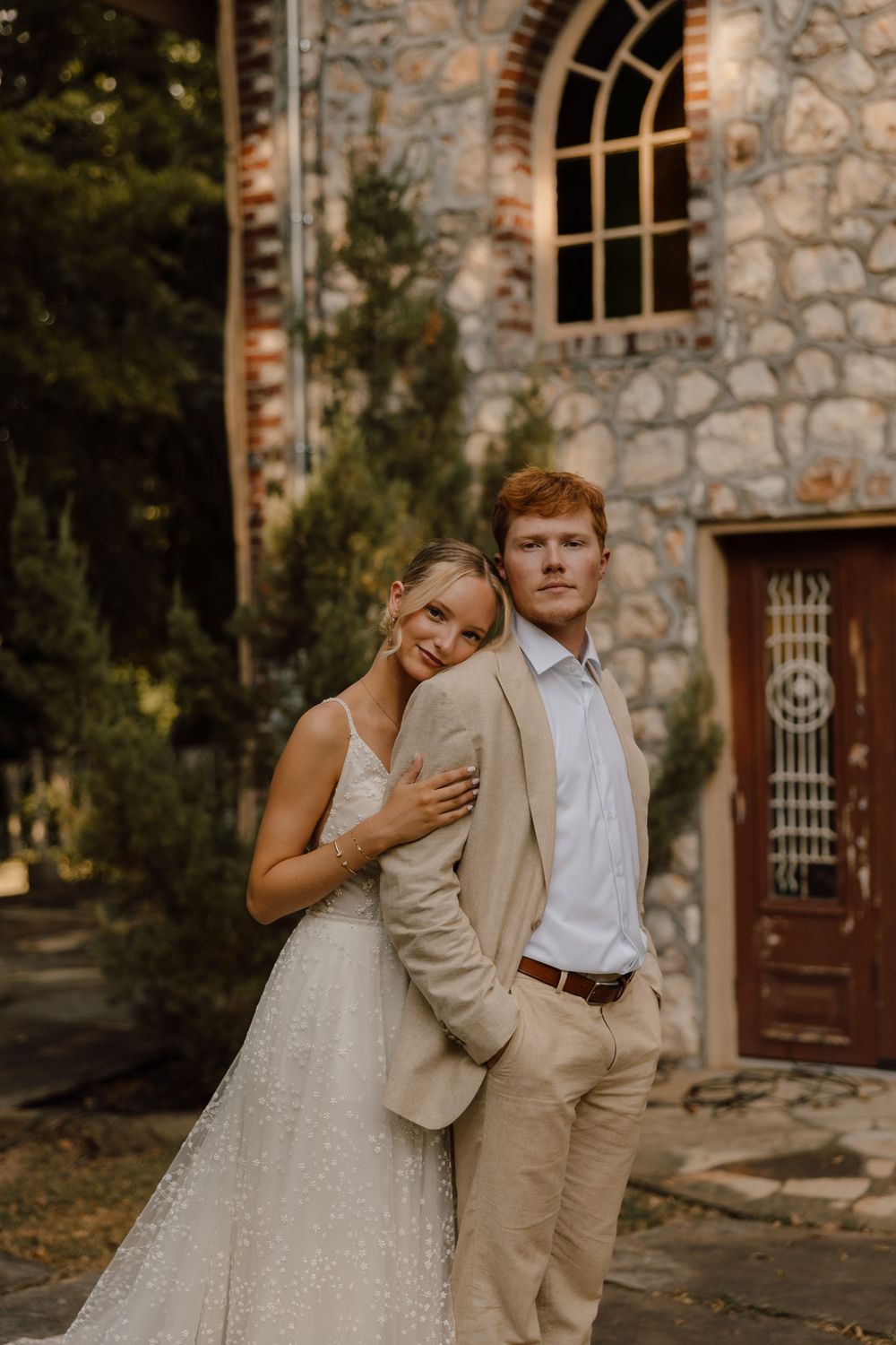 A couple stands lovingly together in front of a rustic stone church with an arched window during a golden hour photoshoot.