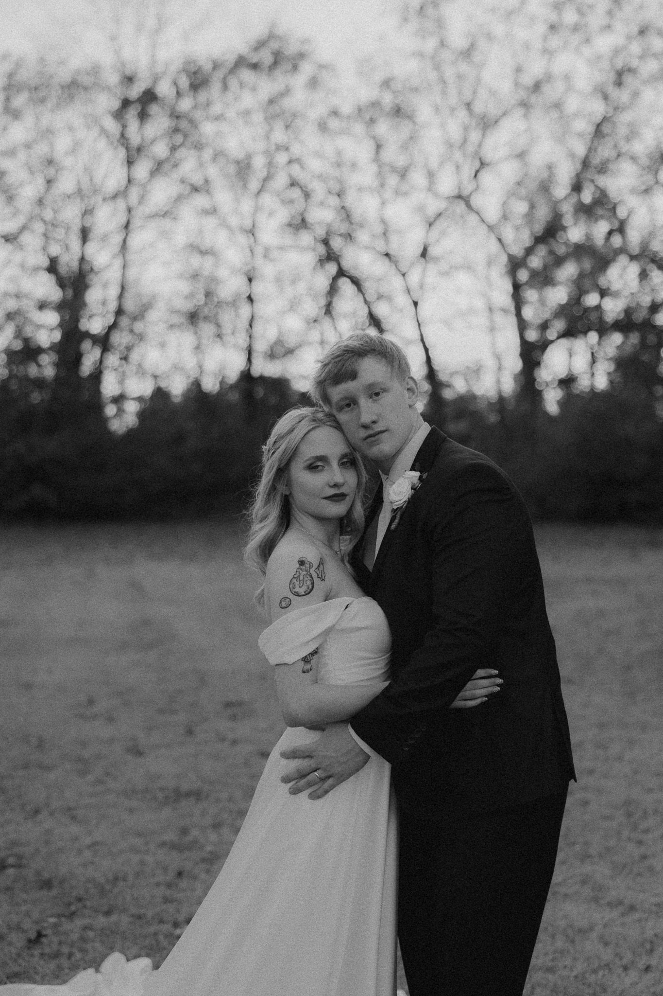 A romantic black and white wedding photo of a couple embracing in a natural outdoor setting with trees in background.
