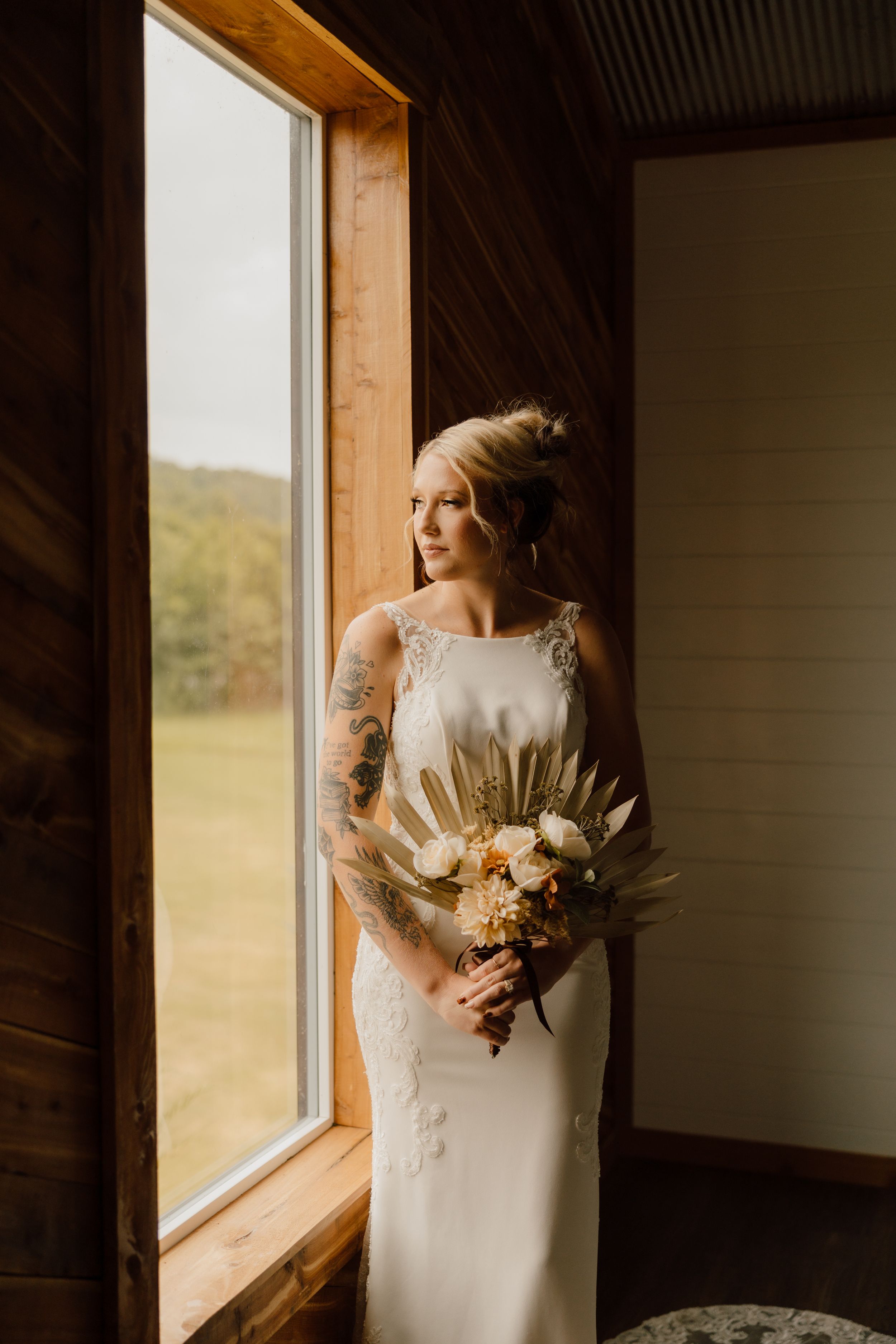 A bride in an elegant white gown stands by a window holding a dried palm leaf and floral bouquet.