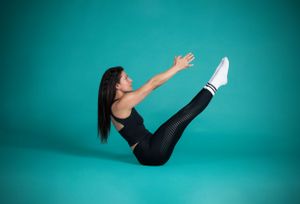 Athletic person in black workout attire performing a v-sit core exercise on turquoise background.