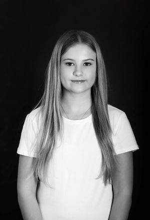 Black and white portrait of a young person with long straight hair wearing a white t-shirt against a dark background.