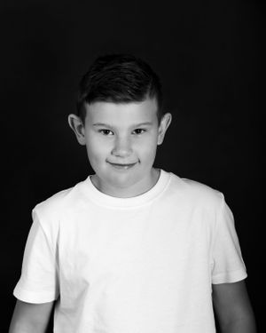 Black and white portrait of a young child in a white t-shirt against a dark background, showing a slight smile.