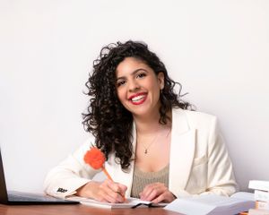 Professional wearing a white blazer and gray top writes with red pen on document while smiling at desk.