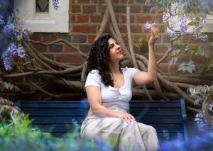 Buisness woman with curly hair sits on a blue bench in a garden surrounded by delicate purple wisteria blooms.