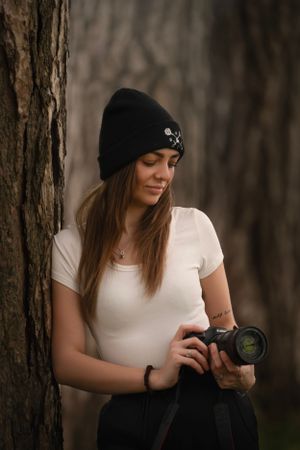 A photographer in a white t-shirt and black beanie holds a camera while leaning against a tree in moody lighting.