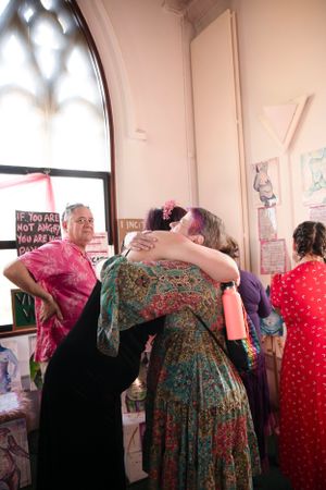 People share an emotional embrace at a gathering in a room with arched windows and decorative posters on the walls.