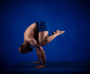 A skilled gymnast performs a handstand balance pose with bent legs against a deep blue studio background.