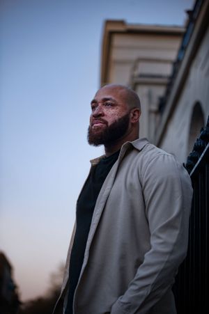 Man with beard wearing black sweater and light jacket stands outdoors at dusk looking into distance.