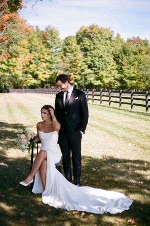 A romantic outdoor wedding photo with a couple posing on a path with autumn trees and wooden fencing in the background.
