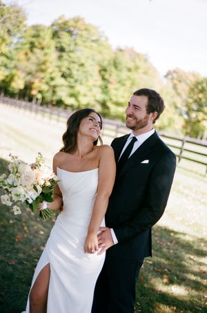 A couple shares a joyful moment outdoors on their wedding day, with sunlit trees and a wooden fence in the background.