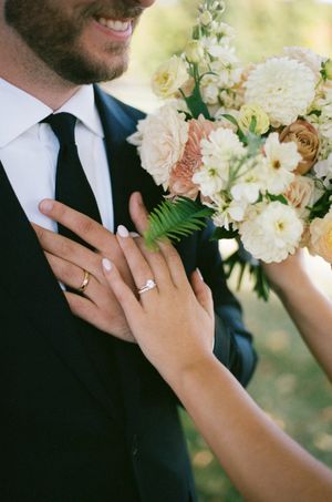 A hand adorned with an engagement ring rests on navy suit next to a romantic bouquet of white and blush flowers.