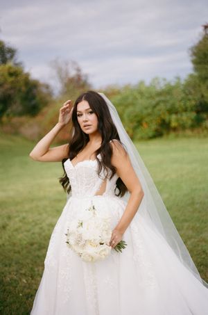 Elegant bride in white lace wedding gown with long veil holds white floral bouquet while posing in outdoor garden setting.