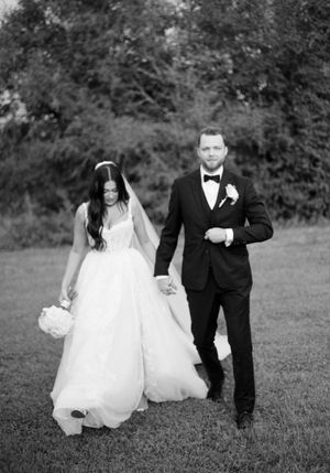 A couple walks hand in hand across a grassy field during their black and white wedding photoshoot.