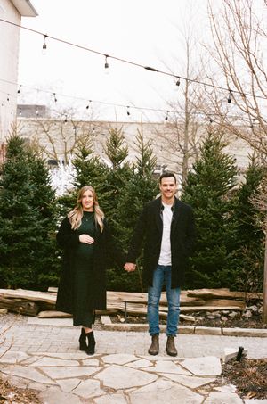 A couple stands on stone pavers in front of evergreen trees with string lights overhead in a vintage-style photo.
