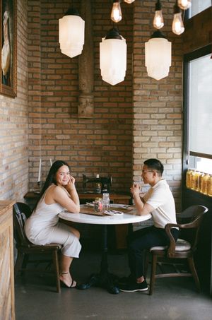 Two people enjoy drinks while sitting at a round table in a cozy cafe with exposed brick walls and pendant lighting.