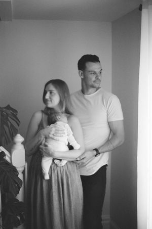 A black and white photograph showing a couple holding a baby in a white outfit against a simple room interior.