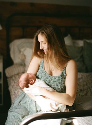 A tender moment as a woman in a gray dress holds and looks down at a swaddled newborn in a dimly lit bedroom.