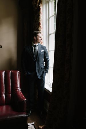 A person in a dark suit stands by a window in a moody room with red leather seating.