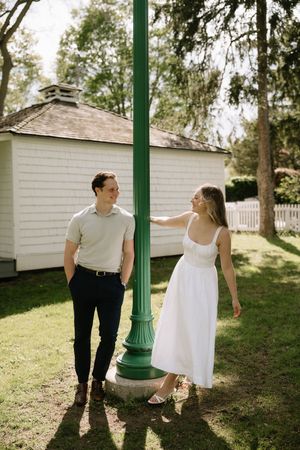 A couple shares a romantic moment by a green lamp post on a sunny day near a white outbuilding.