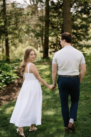 A couple in wedding attire walks hand in hand through a sunlit wooded area during a romantic outdoor photoshoot.