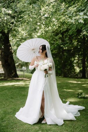 A bride in a flowing white wedding dress poses with a delicate white parasol in a lush garden setting.