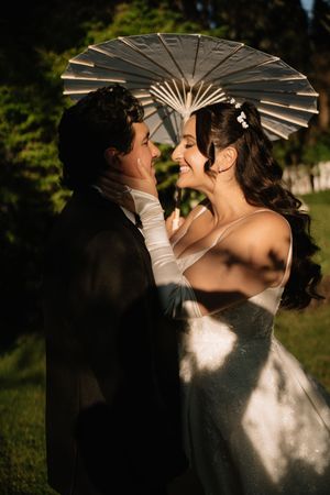 A romantic couple shares a tender kiss under a white parasol in a garden setting with beautiful natural lighting.
