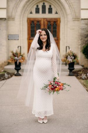 An elegant bride in a white lace dress and veil stands outside a gothic church entrance holding a vibrant floral bouquet.