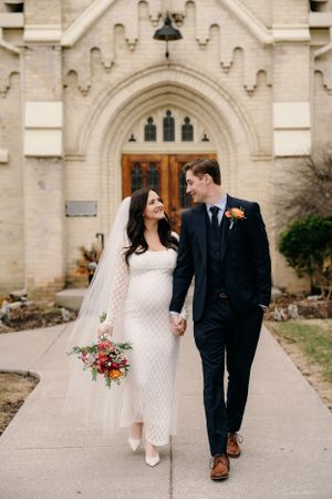 A newlywed couple walking together on a church pathway, with bride in white dress and groom in navy suit.