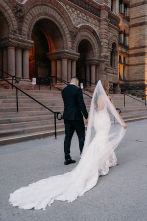 A bride in a flowing white veil and groom ascend the stone steps of a historic church with ornate arched entrances.
