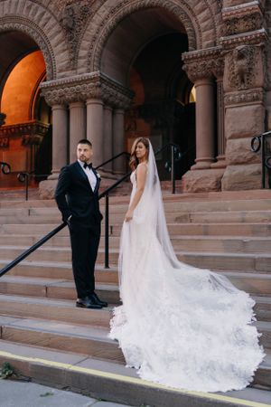 A couple in formal wedding attire poses on grand stone steps of a historic building with ornate archways and columns.