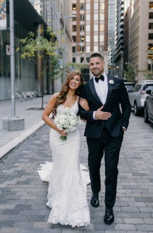 A wedding couple walks together on a city street with tall buildings in the background and a beautiful white bouquet.