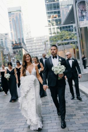 A wedding party walks down a city street with bridesmaids in black dresses following behind the couple.