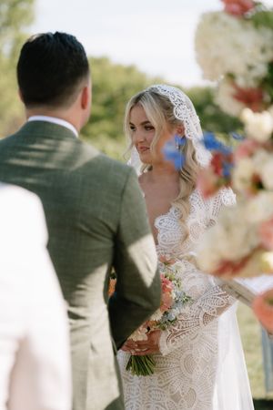 A couple exchanges vows during an outdoor wedding ceremony with the bride wearing an intricate lace gown and veil.