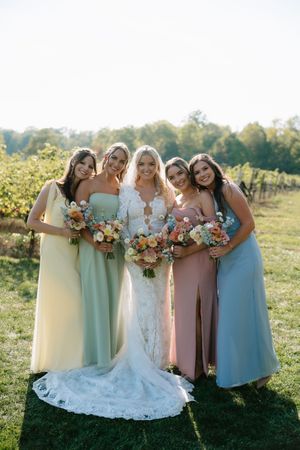 Wedding party in pastel dresses poses together in a sunlit field holding colorful autumn bouquets.