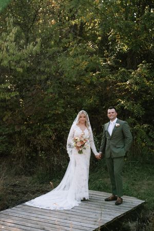 A couple in wedding attire poses together on a wooden deck surrounded by lush green foliage.