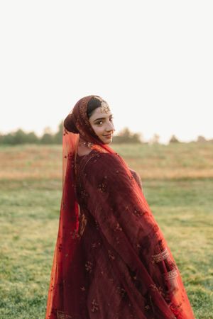 A person wearing a deep red traditional sari with delicate embroidery stands in a grassy field at sunset.