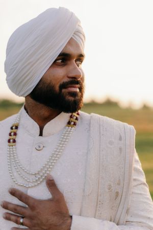 Traditional Indian groom wearing white sherwani with pearl necklaces and turban during sunset outdoors.