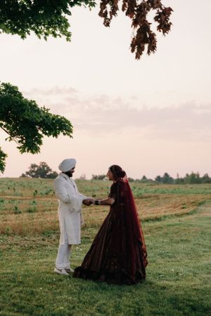 Two people in traditional attire stand together in a green field at sunset, one in white and one in a deep red gown.