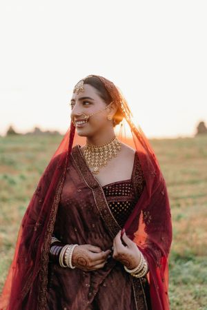 Traditional Indian bridal attire featuring burgundy velvet dress with gold jewelry captured during golden hour sunset outdoors.