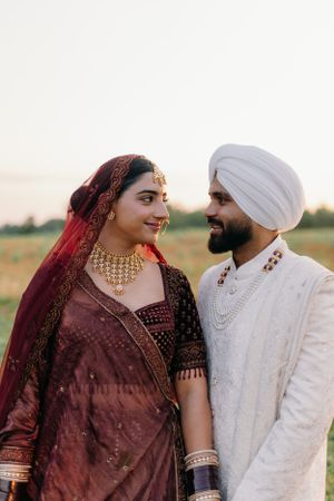 A couple in traditional Indian wedding attire, with the bride in maroon and groom in white, share an intimate moment outdoors.