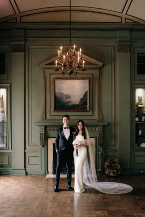 Elegant couple stands in formal attire beneath an ornate chandelier in a room with sage green painted walls and wood floors.