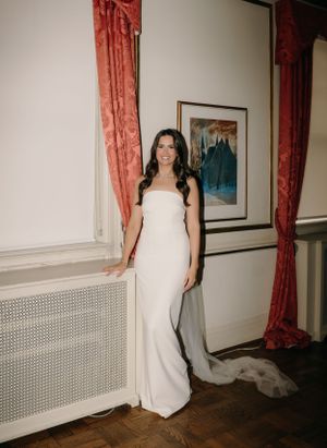 A person in an elegant white strapless wedding gown stands beside red curtains in an elegant room with hardwood floors.