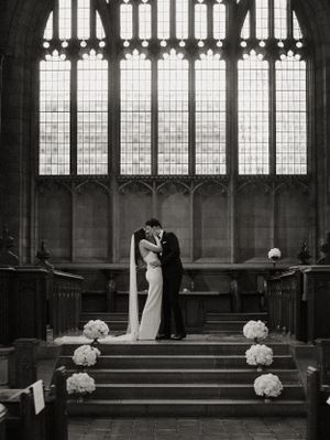 Black and white image of a romantic wedding ceremony in a gothic church with arched windows and floral arrangements on steps.