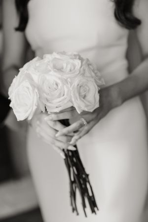 Black and white closeup of hands holding an elegant white rose bridal bouquet against a soft focus background.