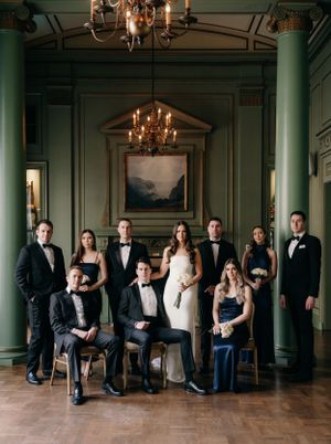 Wedding party stands elegantly in formal attire inside luxurious room with green walls, chandeliers and ornate decor.
