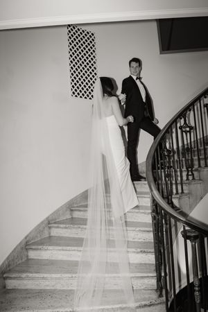 A black and white wedding photo showing a couple elegantly posed on a curved staircase with ornate railings.